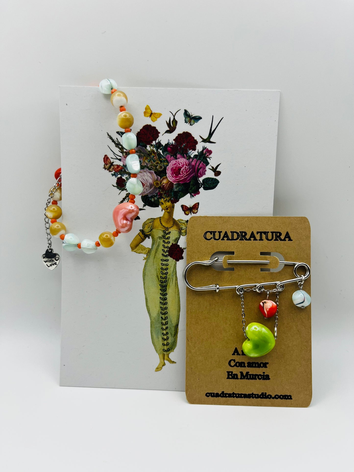 Conjunto artesanal Cuadratura: pulsera Calma con howlita, coral y corazón de cerámica rosa, junto a broche imperdible plateado con colgantes de cerámica verde y rojo, montados a mano en Murcia
