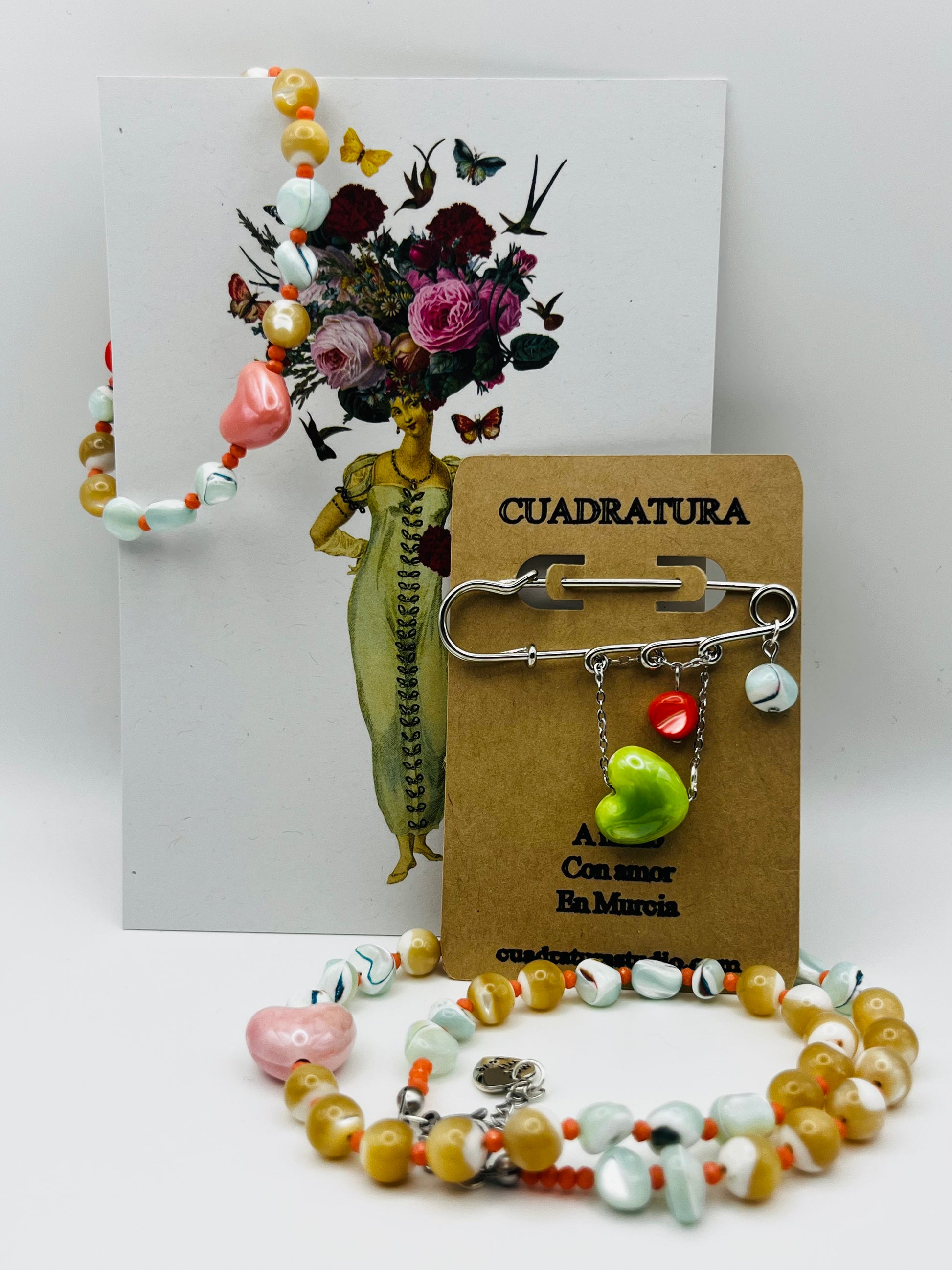 Conjunto de pulsera Calma, collar y broche Calma de Cuadratura Studio: pulsera y collar con cuentas de cerámica, howlita y coral, con corazón rosa; broche imperdible de acero inoxidable con colgantes de howlita y cerámica, sobre tarjeta kraft.