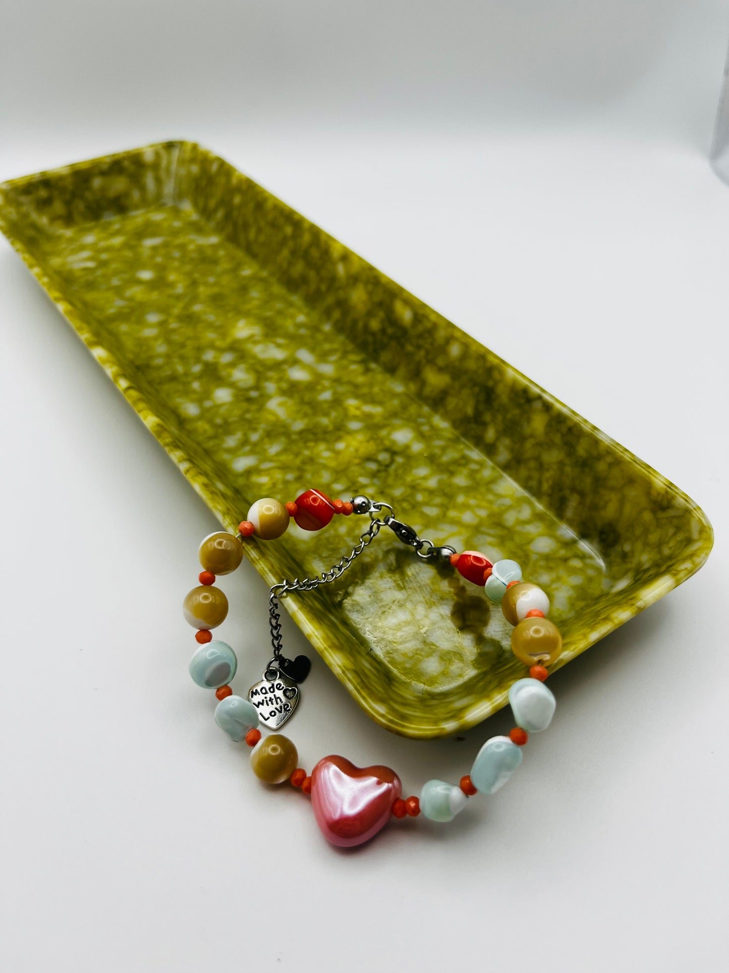 Pulsera artesanal Calma con howlita verde menta, coral rojo y naranja, howlita mostaza y corazón de cerámica rosa, apoyada en bandeja de cerámica verde oliva, montada a mano en Murcia
