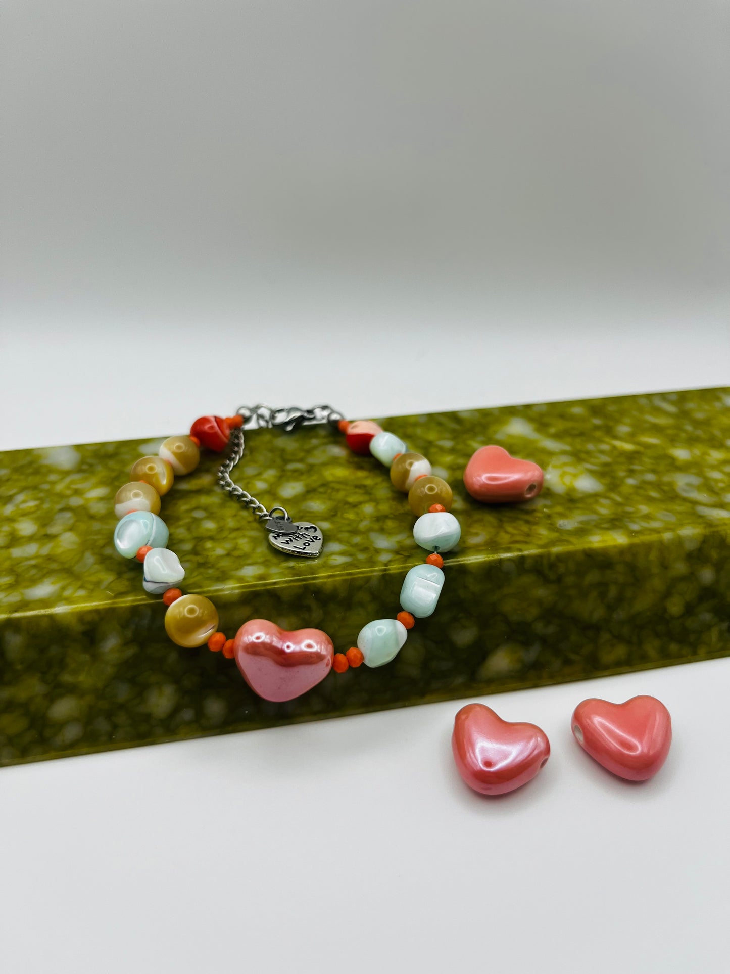 Detalle de pulsera artesanal Calma con howlita verde menta, coral naranja, howlita mostaza y corazón de cerámica rosa, sobre bandeja verde oliva con corazones sueltos, montada a mano en Murcia
